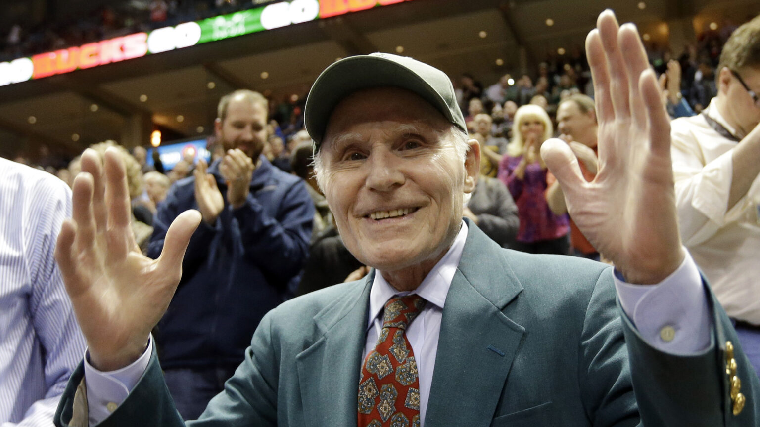 Herb Kohl holds both hands up in the air while standing in the bleachers of an arena, with people standing behind him at progressively higher levels applauding and cheering.