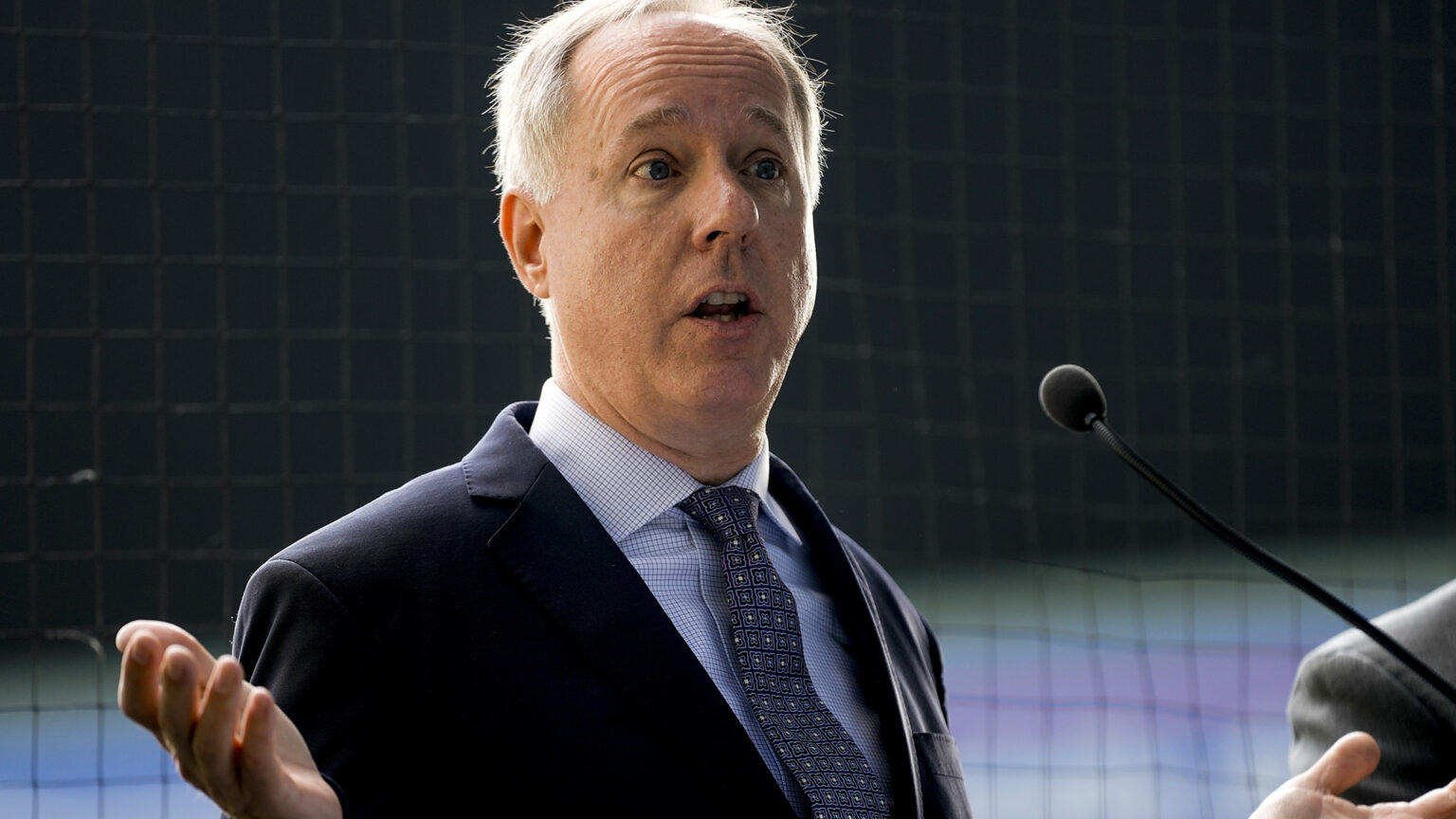 Robin Vos gestures with both hands while speaking into a microphone, with baseball backstop netting behind him.