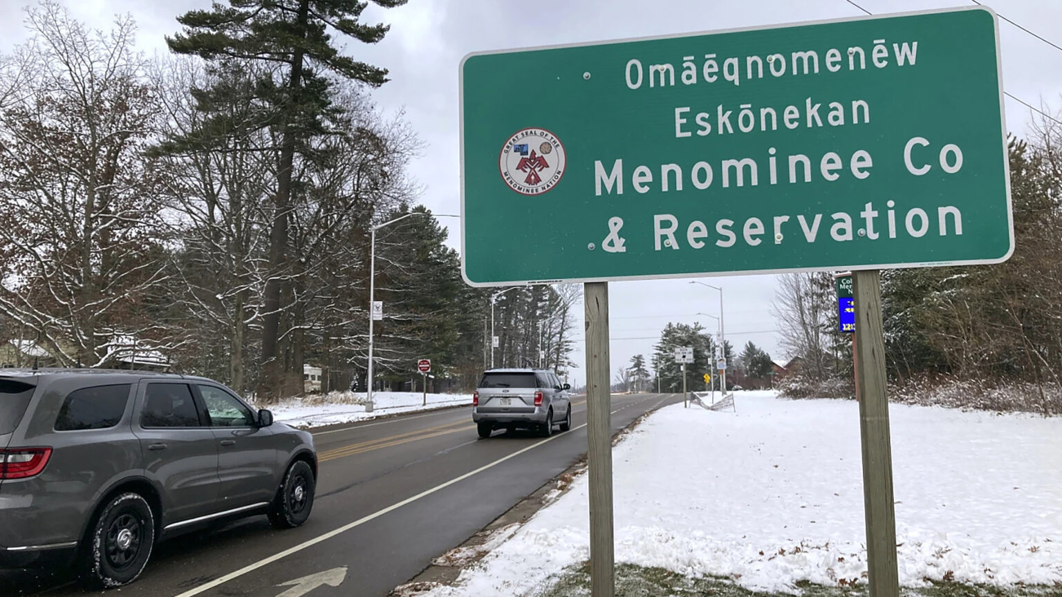 A road sign with the words Omāēqnomenēw Eskōnekan and Menominee Co & Reservation along with the Great Seal of the Menominee Nation stands on wooden posts  in a snow-covered field next to a road with two cars driving past it, with street lights, other signs and trees in the background.