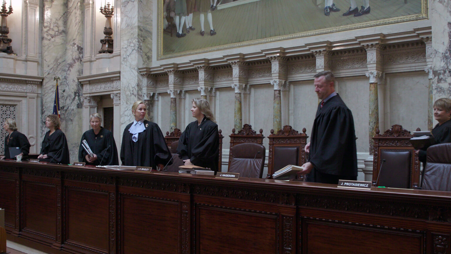 From left to right, Jill Karofsky, Rebecca Dallet, Ann Walsh Bradley Annette Ziegler, Rebecca Bradley, Brian Hagedorn and Janet Protasiewicz stand at a judicial dais amid a row of high-backed leather chairs, with another row of high-backed wood and leather chairs behind them, in a room with marble masonry, a large painting and a U.S. flag.