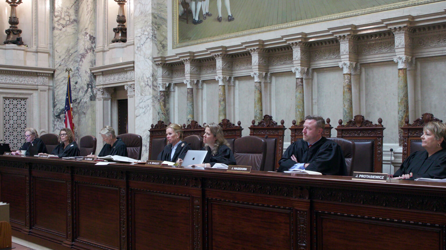From left to right, Jill Karofsky, Rebecca Dallet, Ann Walsh Bradley Annette Ziegler, Rebecca Bradley, Brian Hagedorn and Janet Protasiewicz sit at a judicial dais in a row of high-backed leather chairs behind them, with another row of high-backed wood and leather chairs behind them, in a room with marble masonry, a U.S. flag and a large painting.