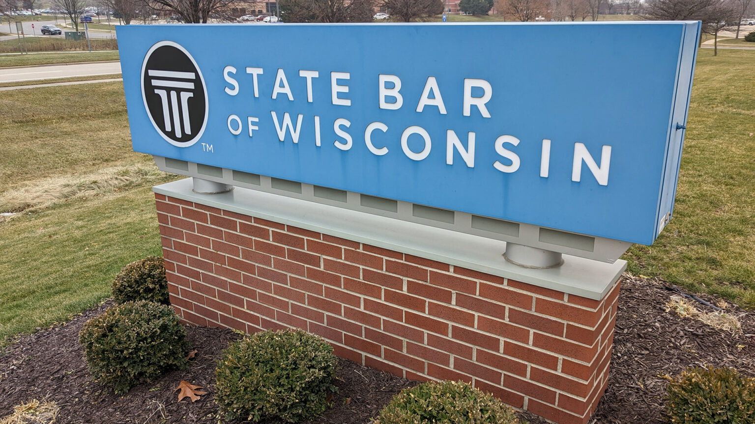 A sign with a stylized logo of a Doric column and the words State Bar of Wisconsin stands on a brick pedestal surrounded by small bushes and mulch in a lawn, with roads, sidewalks and leafless trees in the background.
