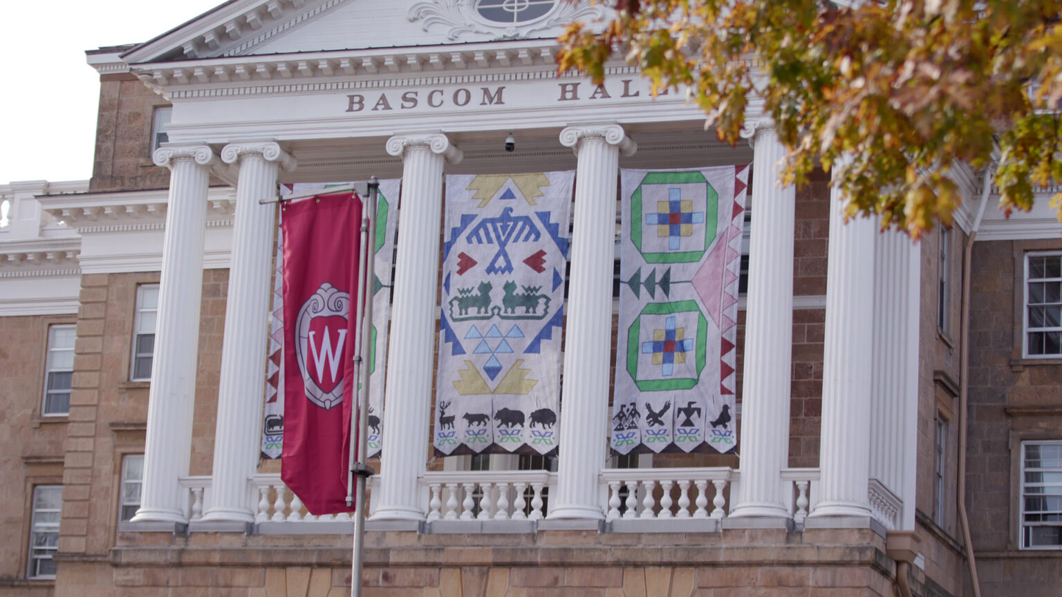 Three banners with different patterns are displayed in between Ionic pillars supporting a pediment at the front of a multi-story building with masonry walls, multi-paned glass windows, with the words Bascom Hall on its face, and with a banner with the UW-Madison crest logo and out-of-focus leaves in the foreground.