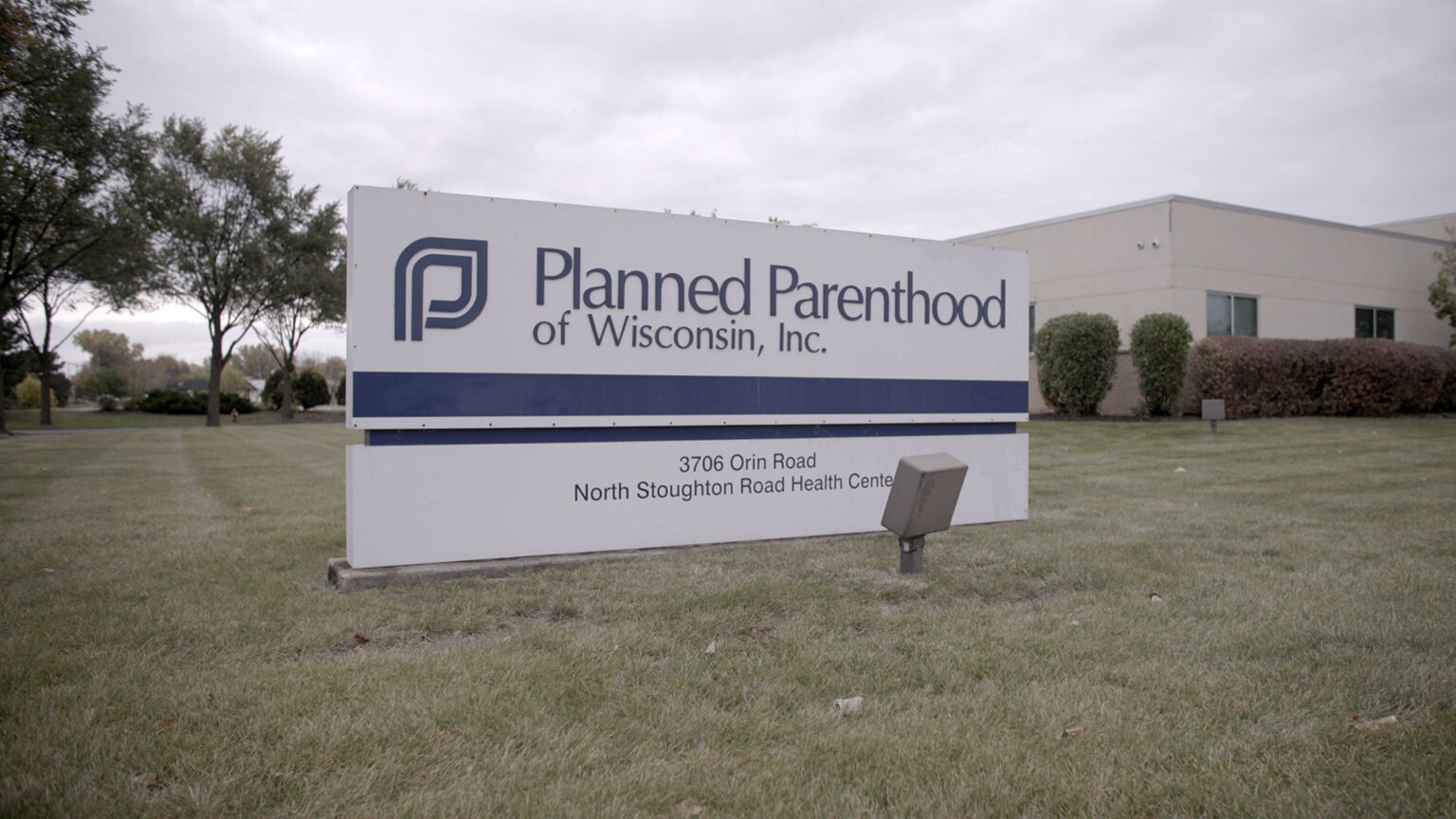 A sign reading Planned Parenthood of Wisconsin, Inc. with an address stands in front of a single-story building surrounded by a lawn, bushes and trees.