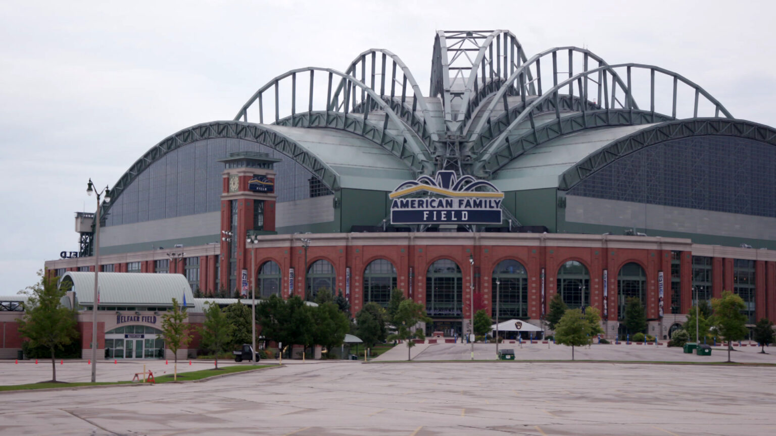 A retractable roof is in a closed position on top of a baseball stadium with a sign over its home plate entrance reading American Family Field, with an empty parking lots and trees in the foreground.