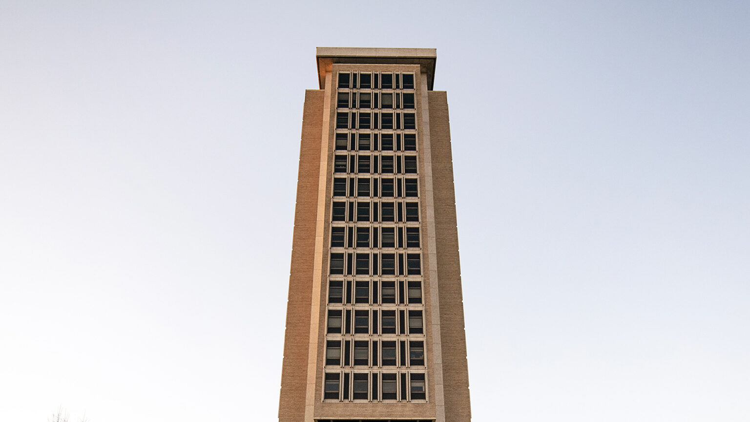 A tower with a brick-and-concrete surface framing 13 stories with four windows apiece stands under a cloudless sky, with sunshine illuminating one side of the building.