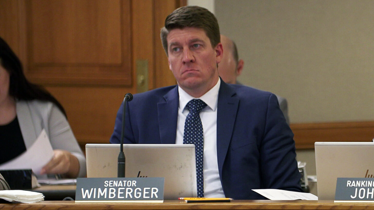 Eric Wimberger sits at a table behind an open laptop computer and microphone, with a nameplate in front of him reading Senator Wimberger, with other people seated in the background  in front of a closed door.