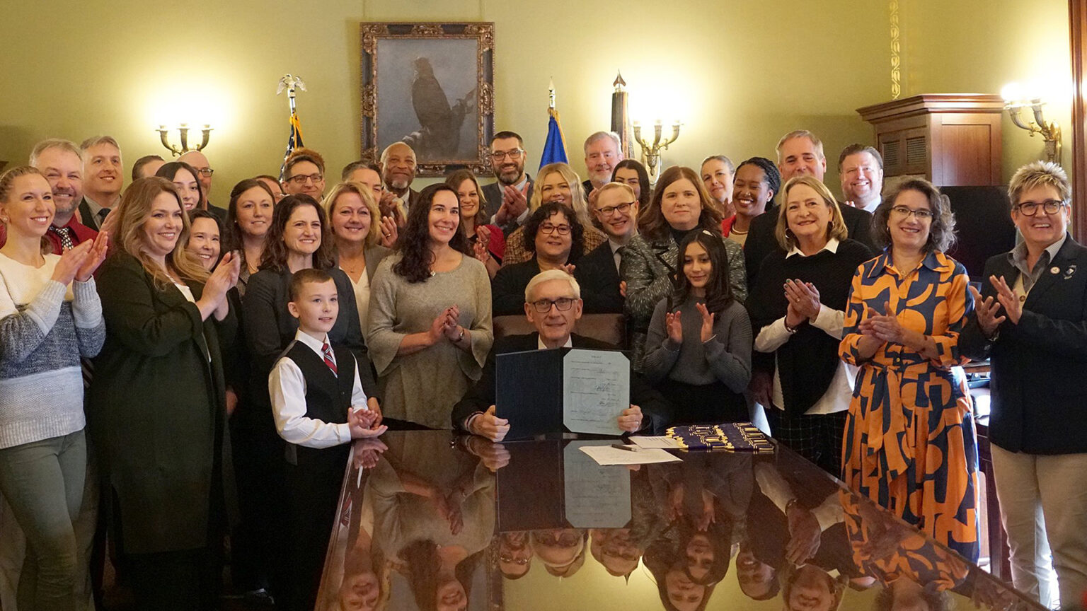 Tony Evers holds an open folder with a document on one side while seated at a table with a glass surface, reflecting people who are standing behind and around him and applauding, in a room with electric wall sconces, the U.S. and Wisconsin flags and a painting.
