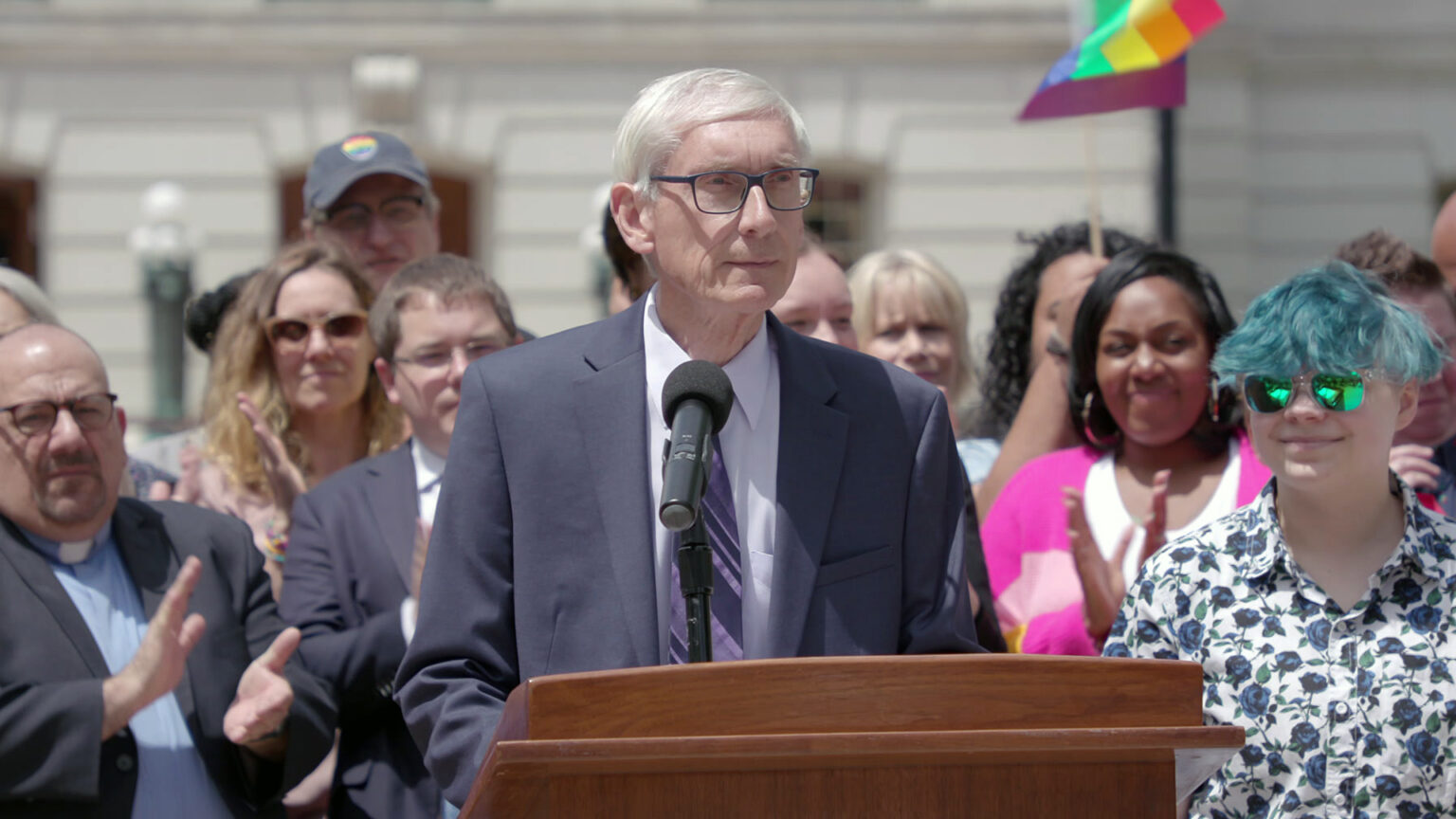 Tony Evers speaks into a microphone while standing behind a wood podium, with other people applauding while standing around and behind him, with an out-of-focus building in the background.