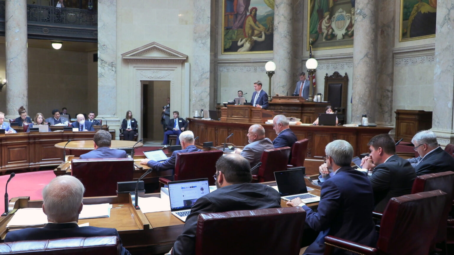 State senators sit at wood desks arranged in a semi-circle and listen to another senator direction the session from a wooden legislative dais, in a room with marble masonry and pillars, large paintings, and a second-story observation gallery.