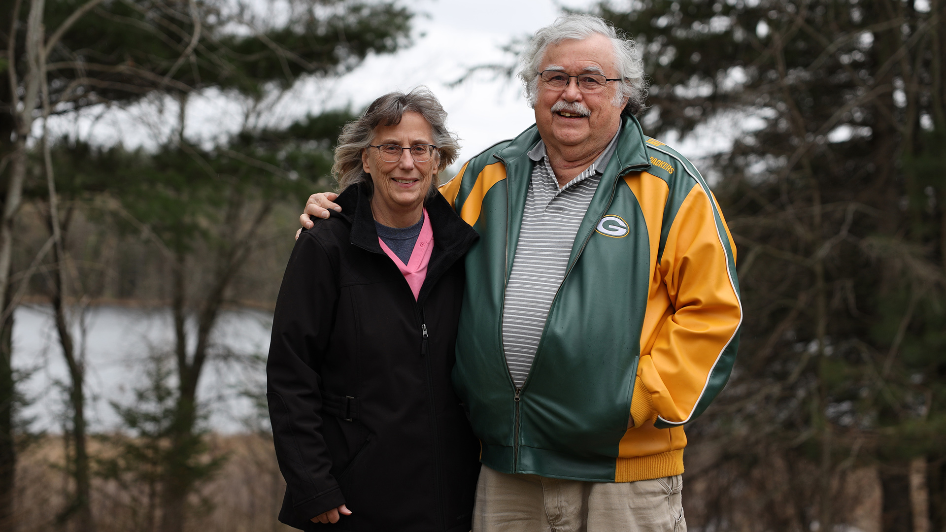Joanne and Howard Pahl pose for a portrait while standing in front of trees and a body of water.