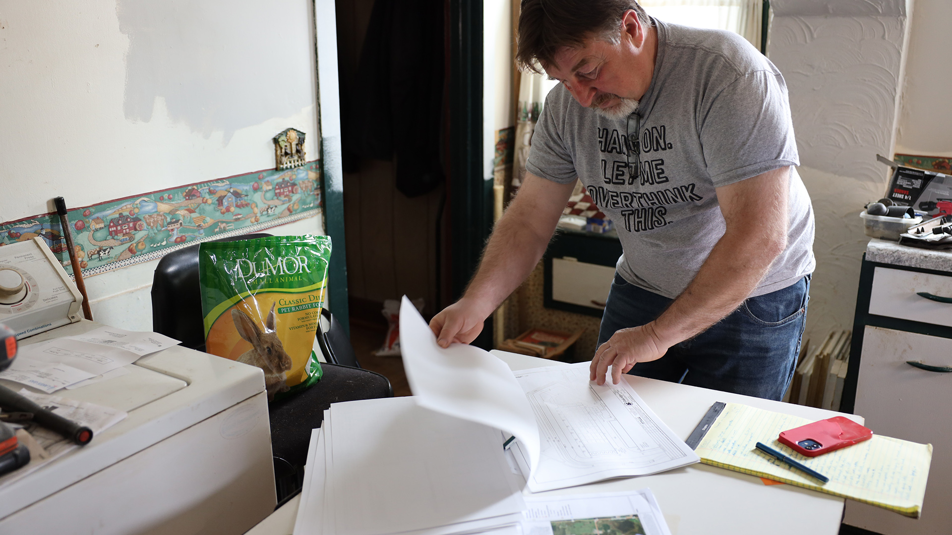 Tim Schmitz stands over a table and pages through documents, with a legal notepad, smart phone and pen on its surface, in a room with a washing machine, cabinets and chairs.