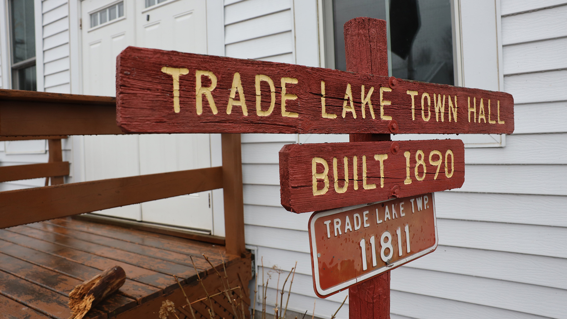 A wood sign with engraved painted letters reads "Trade Lake Town Hall" and "Built 1890" along with a metal address sign reading "Trade Lake Twp. 11811" stands in front of a building with two windows framing double doors in front of a wood ramp.