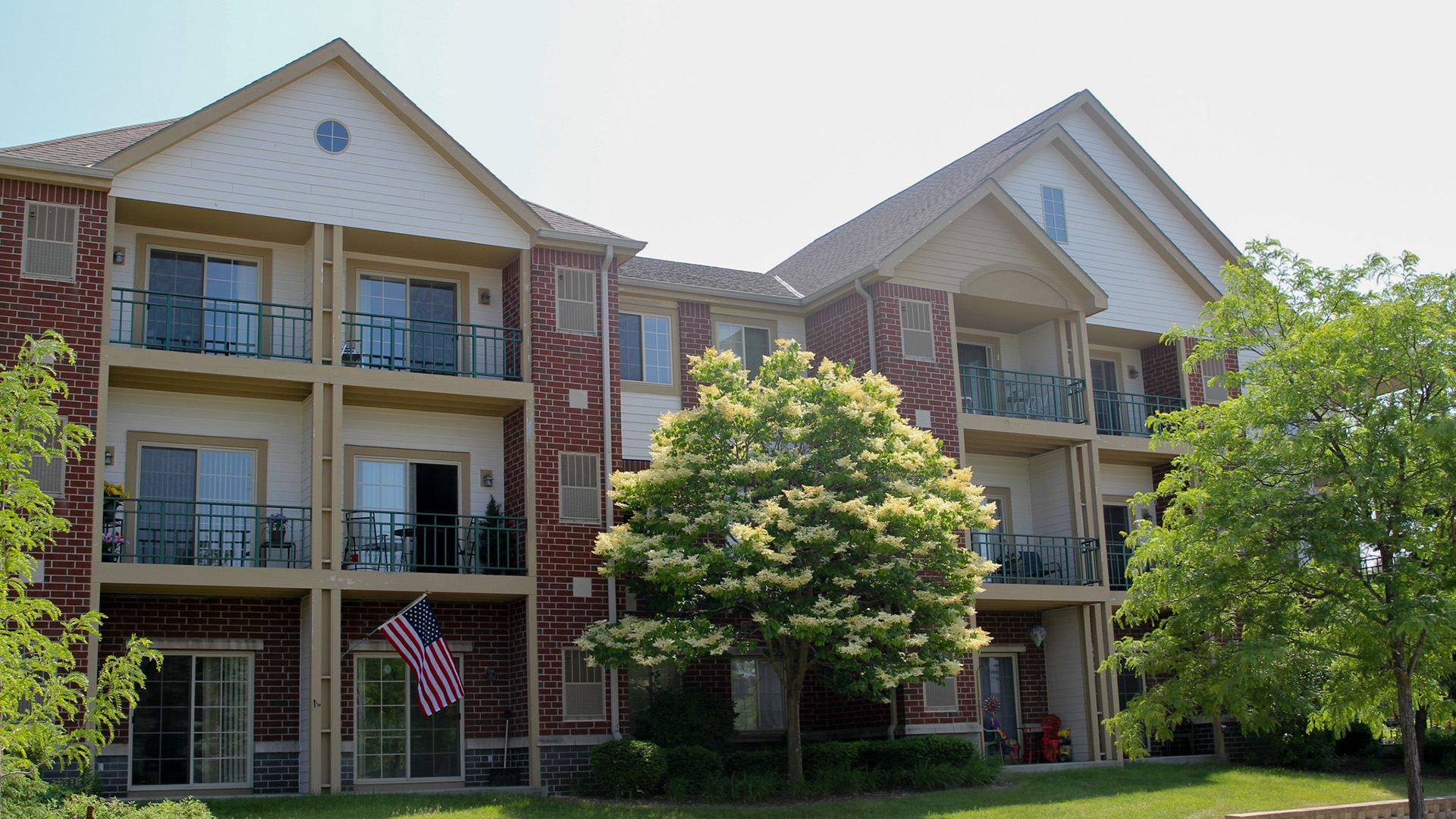 Sunshine illuminates a multi-story residential building with patios and decks alternating with windows, topped by multiple gable roofs, with a U.S. flag mounted on a wall in front of one unit and trees in the foreground.