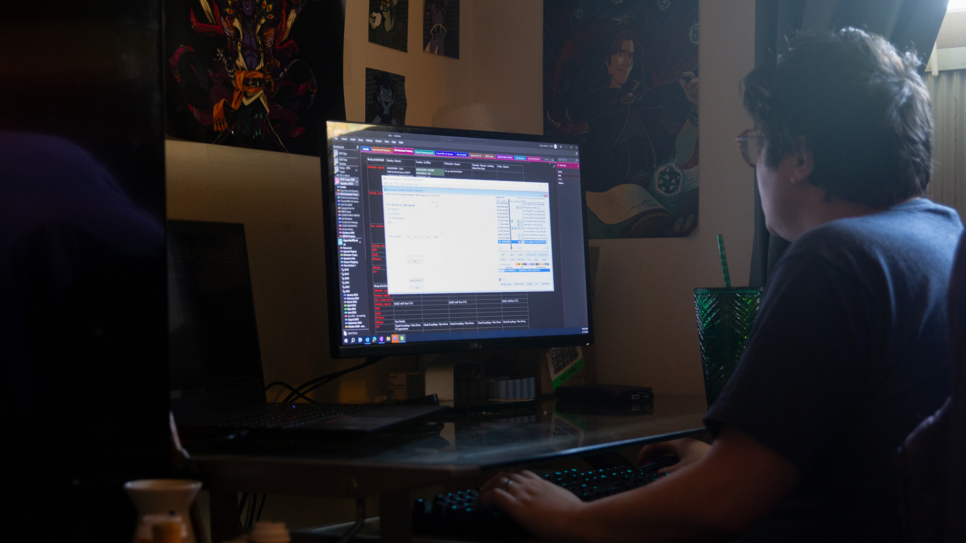 Robin Alexander uses a keyboard while looking at a computer monitor on a desk, in a room with multiple posters on the wall next to a window with open drapes.