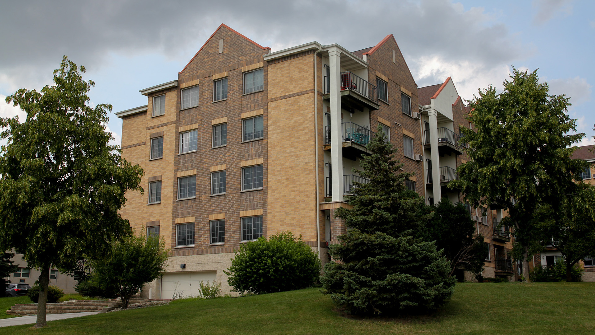A cloud shadows a multi-story residential building with windows placed amid brick siding along one face and multiple exterior decks on another, topped by multiple gabled roofs, with trees on a lawn in the foreground.