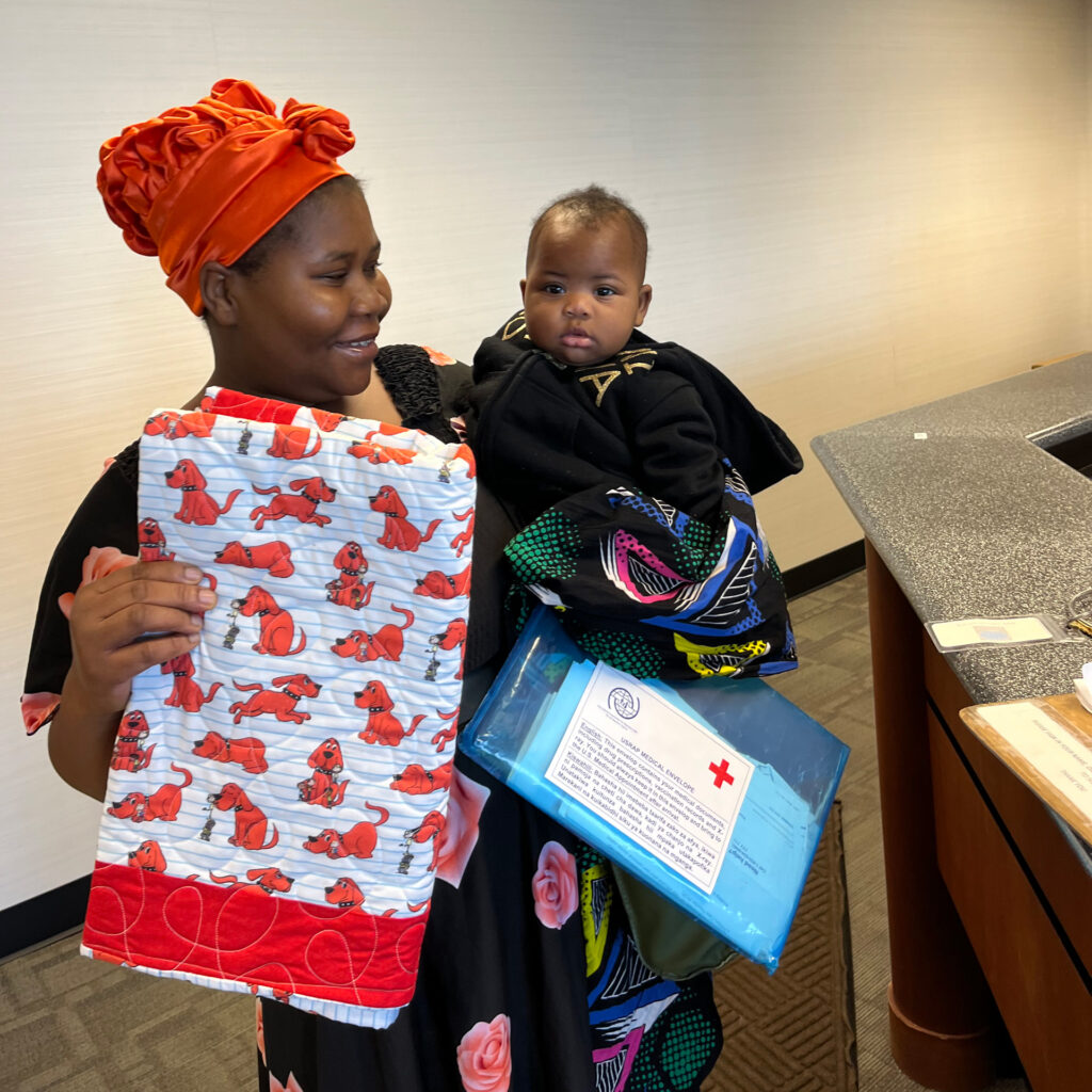 A woman smiles holding a baby, a packet of documents and a quilt with a "Clifford the Big Red Dog" pattern on it.