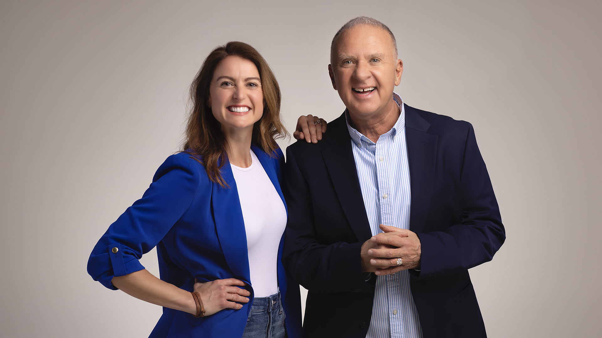 Emmy Fink has her hand on the shoulder of John McGivern, and both wear blazers while standing against a plain gray background and smiling at the camera.