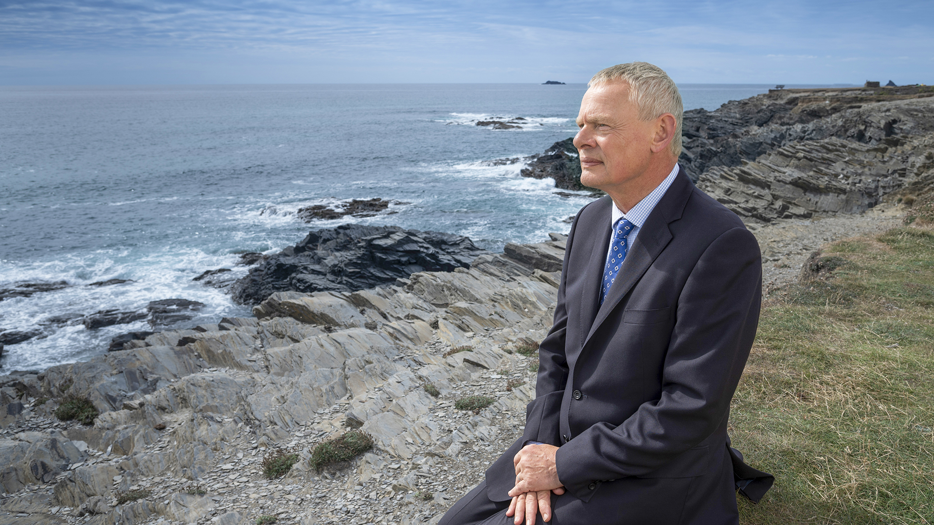 Doc Martin wears a suit and tie while he sits on a grassy hill overlooking a rocky shoreline.