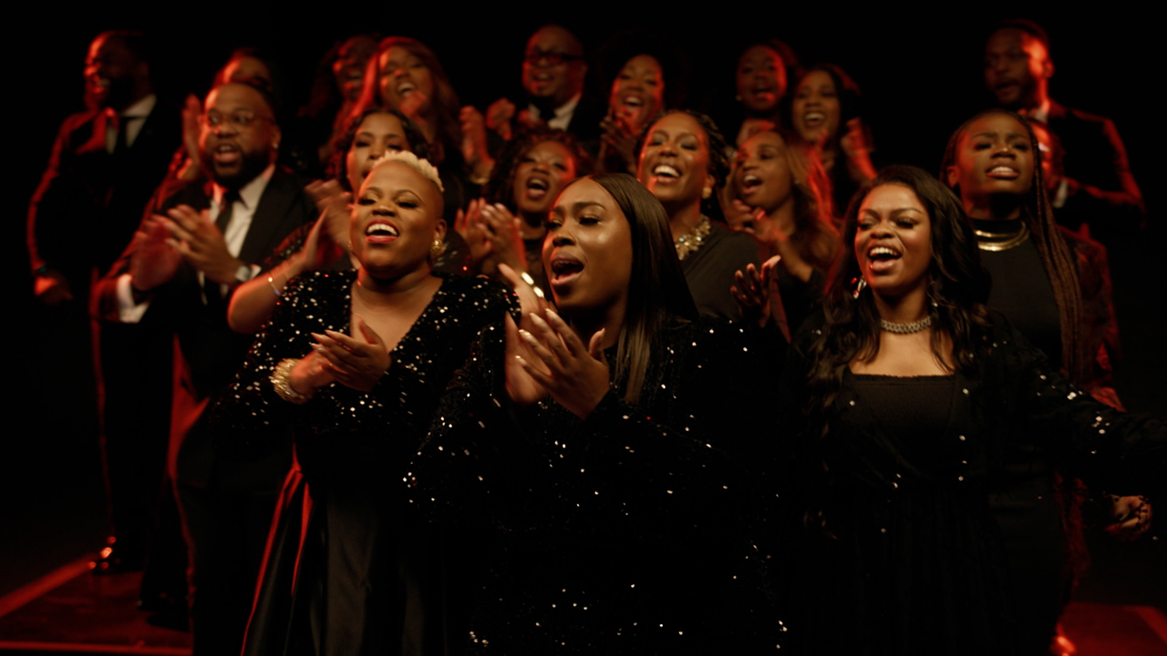 A Black gospel choir dressed in black clothing sings and claps their hands under dim lighting during a performance.