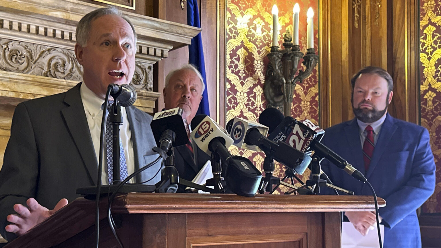 Robin Vos stands and speaks into multiple microphones with the flags of different media organizations mounted to the top of a wood podium, with two other people standing behind him and to his side, in a room with a carved fireplace lintel, the Wisconsin flag, electric wall sconces and toile wallpaper.