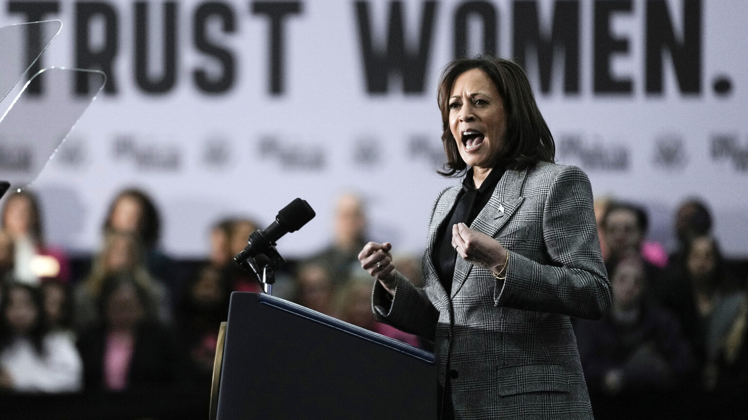 Kamala Harris speaks and gestures with both hands while standing behind a podium with two mounted microphones and facing two teleprompter mirrors, with out-of-focus people standing in the background beneath a sign reading Trust Women.