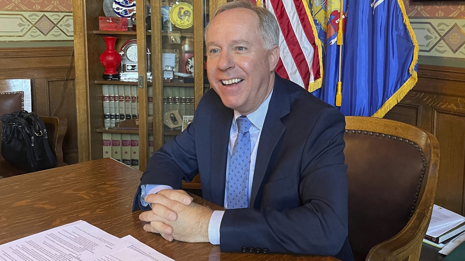 Robin Vos sits in a wood and leather curved back chair and rests folded hands on a wood table with papers on its surface, in a room with wood panels, wall paper, the U.S. and Wisconsin flags, and a cabinet with law books and decorative items.