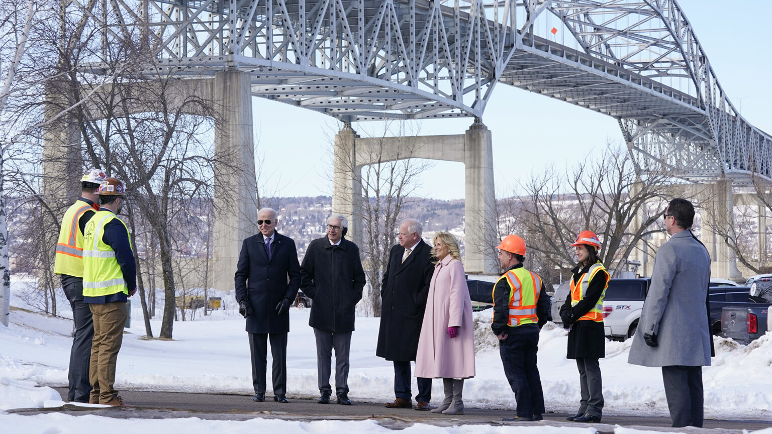 Joe Biden, Tony Evers, Tim Walz and Jill Biden stand alongside two people wearing safety vests and helmets, with three other people facing them on a sidewalk in a snow-covered area with leafless trees and parked vehicles at the base of a bridge with concrete pillars and a steel girder superstructure, with foothills on the horizon.