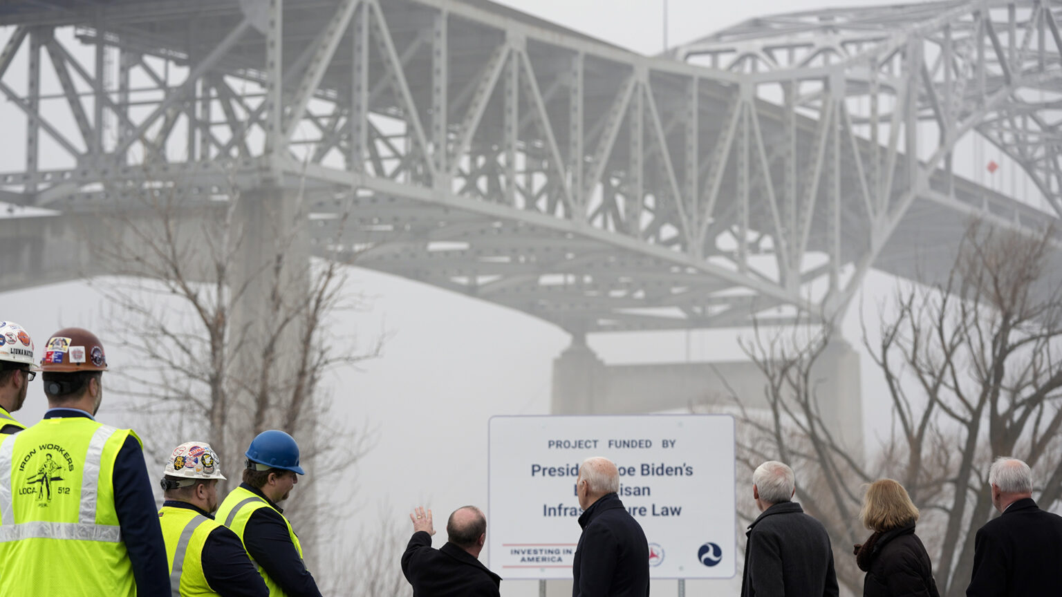Joe Biden stands among four people wearing safety vests and four other people who are facing leafless trees, a sign reading Project Funded by President Joe Biden's Bipartisan Infrastructure Law and the concrete pillars and steel girder superstructure of a bridge partially obscured in fog.