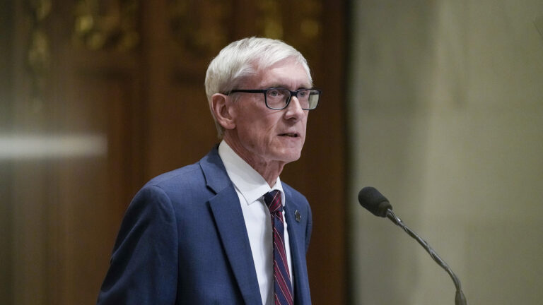Tony Evers speaks into a microphone while standing in a room with wood wall paneling and marble masonry.