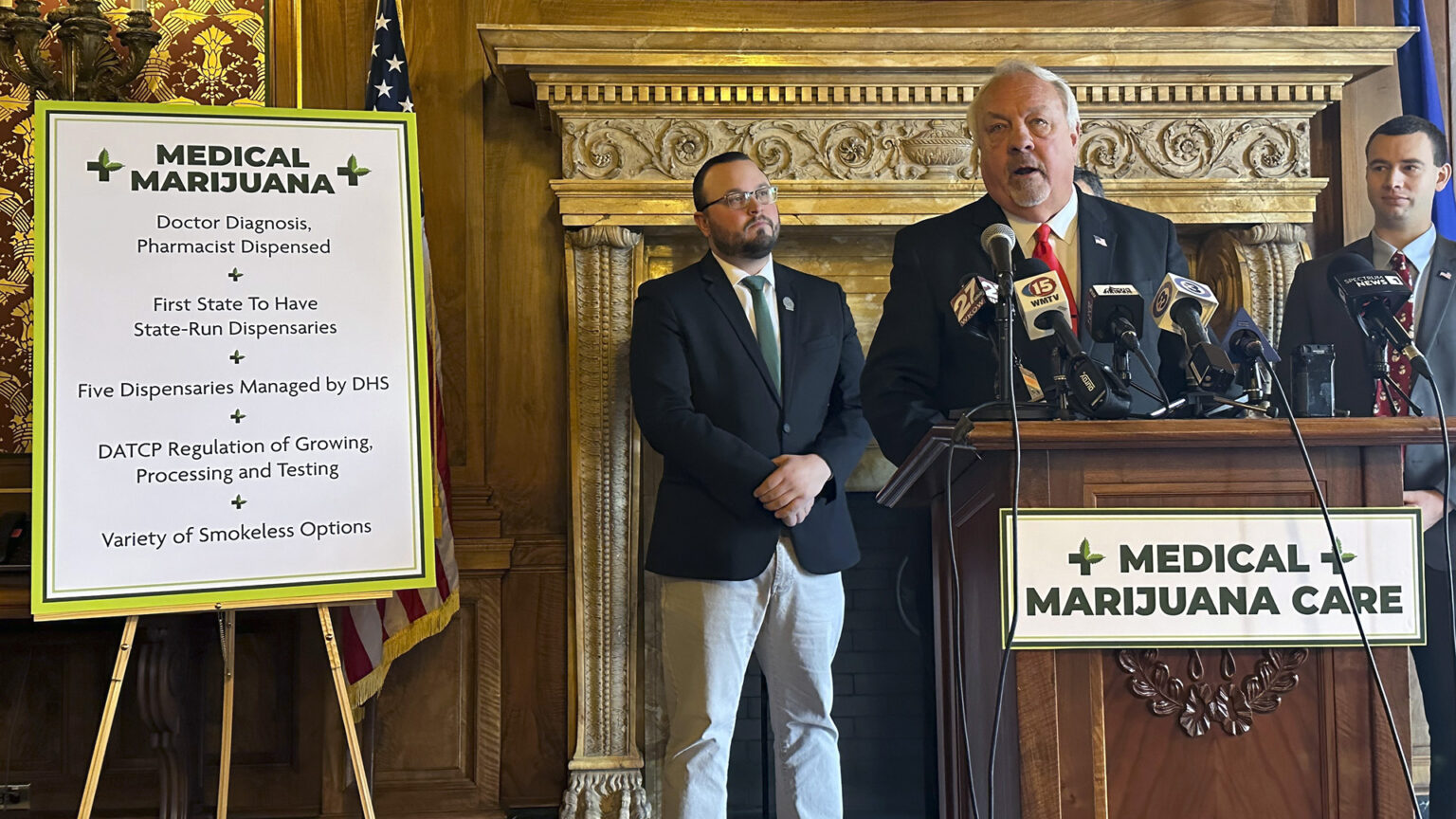 Jon Plumer stands and speaks into multiple microphones mounted to a wood podium with a sign affixed to its front reading Medical Marijuana Care and another sign to his side reading Medical Marijuana, Doctor Diagnosis, Pharmacist Dispensed, First State To Have State-Run Dispensaries, Five Dispensaries Managed by DHS, DATCP Regulation of Growing, Processing and Testing, and Variety of Smokeless Options, with two other people standing behind him in a room with a carved fireplace lintel, the U.S. and Wisconsin flags, and toile wallpaper.