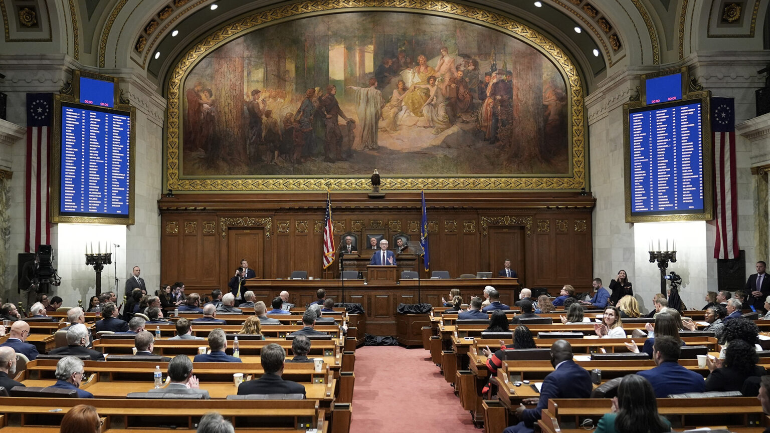 Tony Evers speaks while standing behind a podium on a wood legislative dais, with three people seated behind him on the next level and dozens of people facing him while seated in rows of wood desks, in a high-ceilinged marble masonry room with a taxidermy bald eagle, the U.S. and Wisconsin flags and a large painting with an arched top behind the dais, digital vote registers on either side and U.S. flag bunting beneath two arches on the side wall.