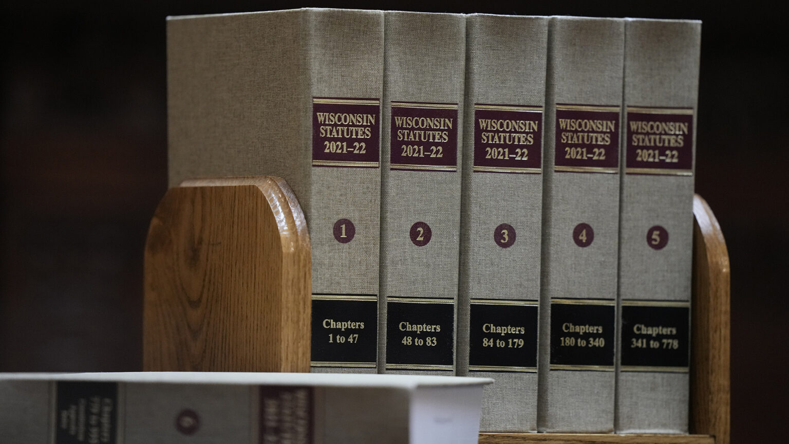 A row of five cloth-bound books with the title Wisconsin Statutes 2021-22, with labels marking the volume number and chapters included stand on a small wood bookshelf, with another book resting on its side and out-of-focus in the foreground.