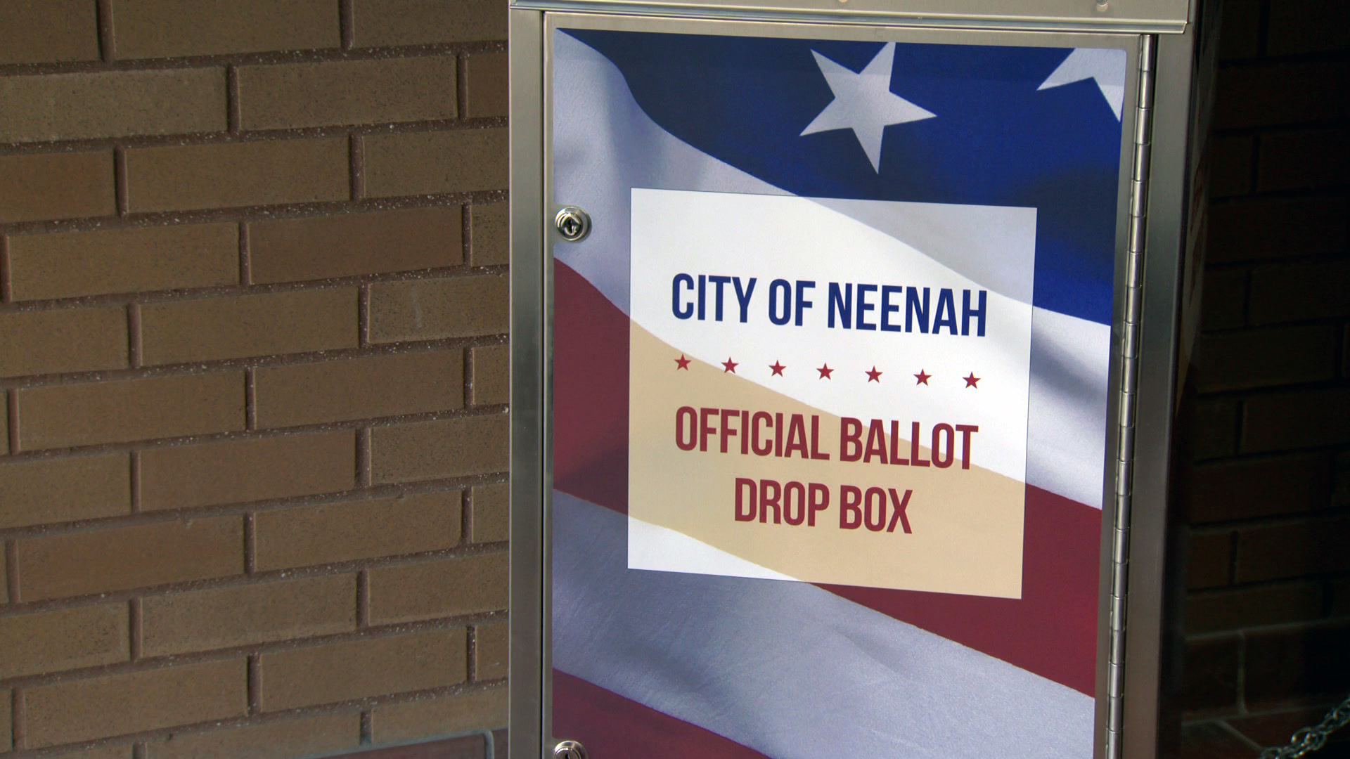 A metal box with a graphic of the U.S. flag with a sign reading "City of Neenah" and "Official Ballot Drop Box" on its front stands in front of a brick wall.