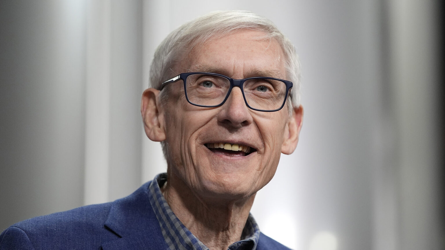 Tony Evers smiles while speaking, with out-of-focus fermentation tanks in the background.