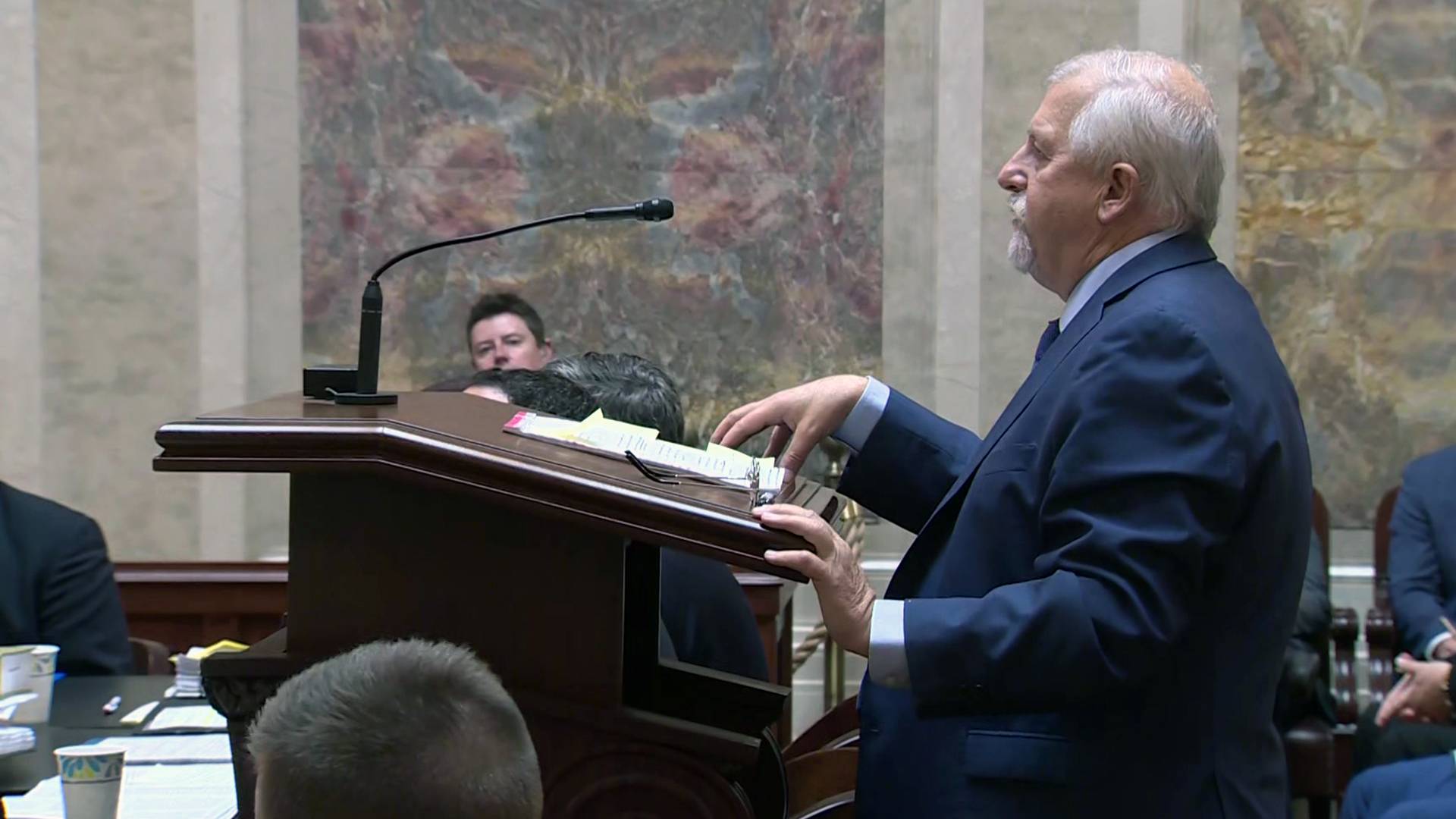 Rick Esenberg gestures with his right hand while standing behind a wood podium and speaking into a microphone mounted to its top with other people seated in the foreground and background in a room with marble walls.