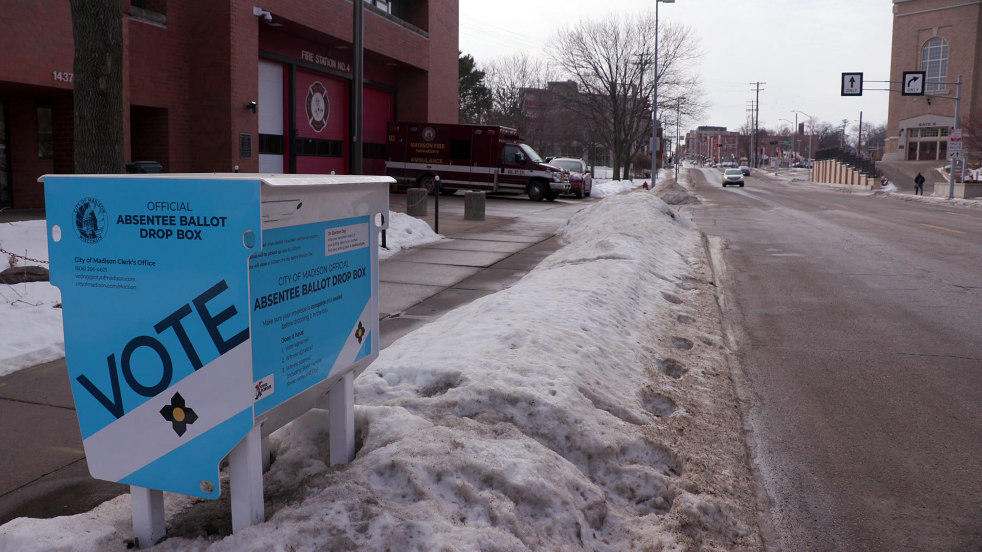 A metal drop box with the word "Vote" stands in a dirty snowbank on the side of a road with a parked ambulance and buildings in the background.