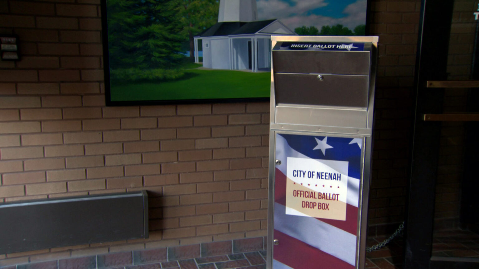 A metal box with a delivery slot at its top and a graphic of the U.S. flag with a sign reading City of Neenah and Official Ballot Drop Box on its front stands in front of a brick wall with a painting of a building and trees.