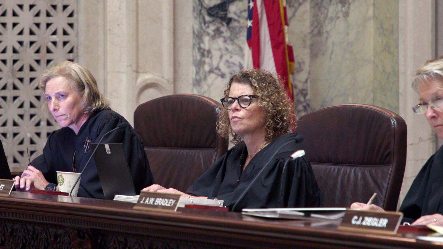 From left to right, Jill Karofsky, Rebecca Dallet, and Ann Walsh Bradley sit at a judicial dais in a row of high-backed leather chairs behind them, in a room with marble masonry and a U.S. flag.