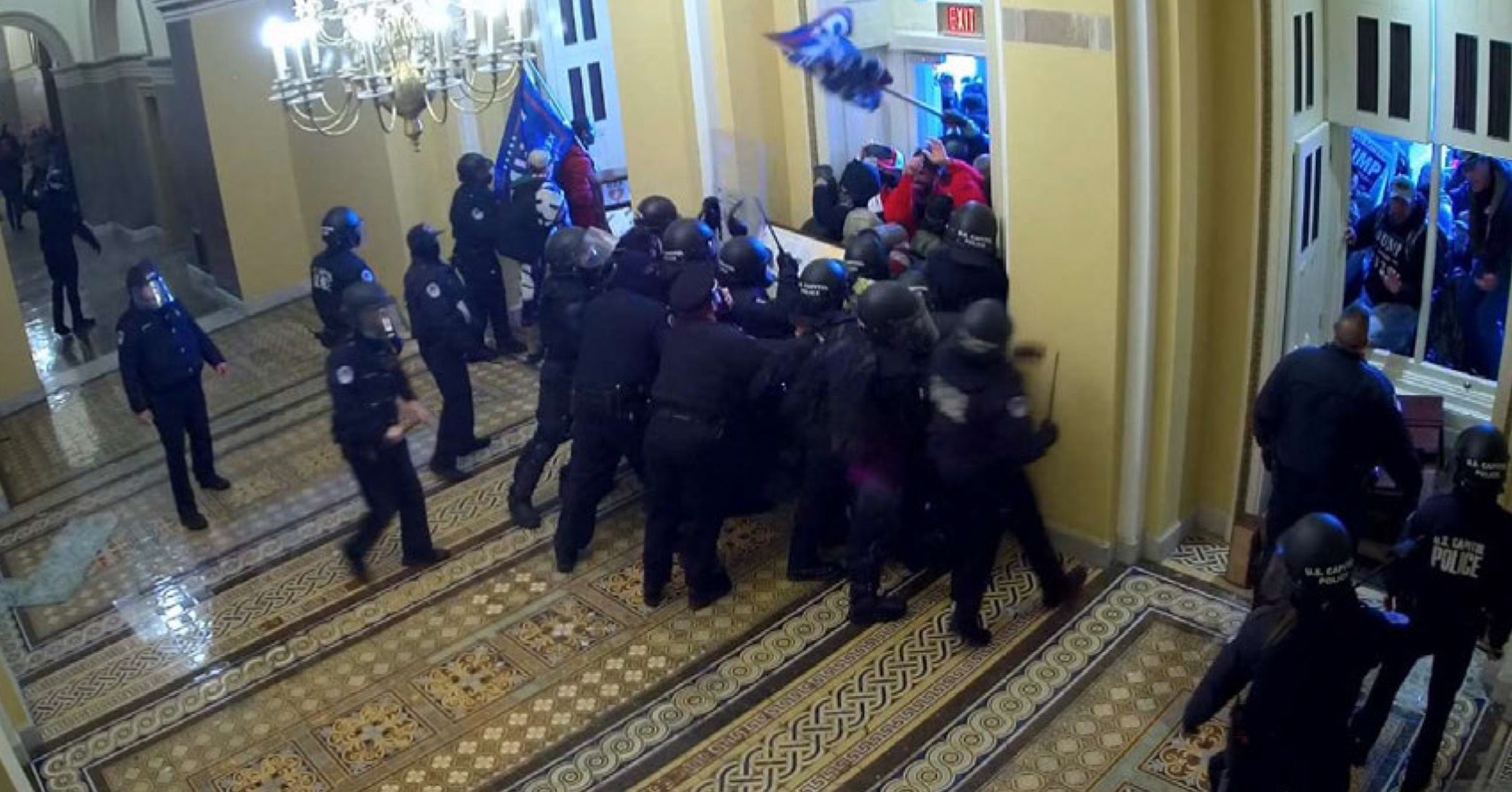 An overhead image shows a group of police wearing helmets and other protective gear, with some carrying batons, standing in a cluster at the entrance to a doorway and facing a crowd of people, some of whom are carrying flags, in a room with a tile floor and illuminated chandelier, with other officers standing in front of windows windows with no glass in the frames on one side of the entry.