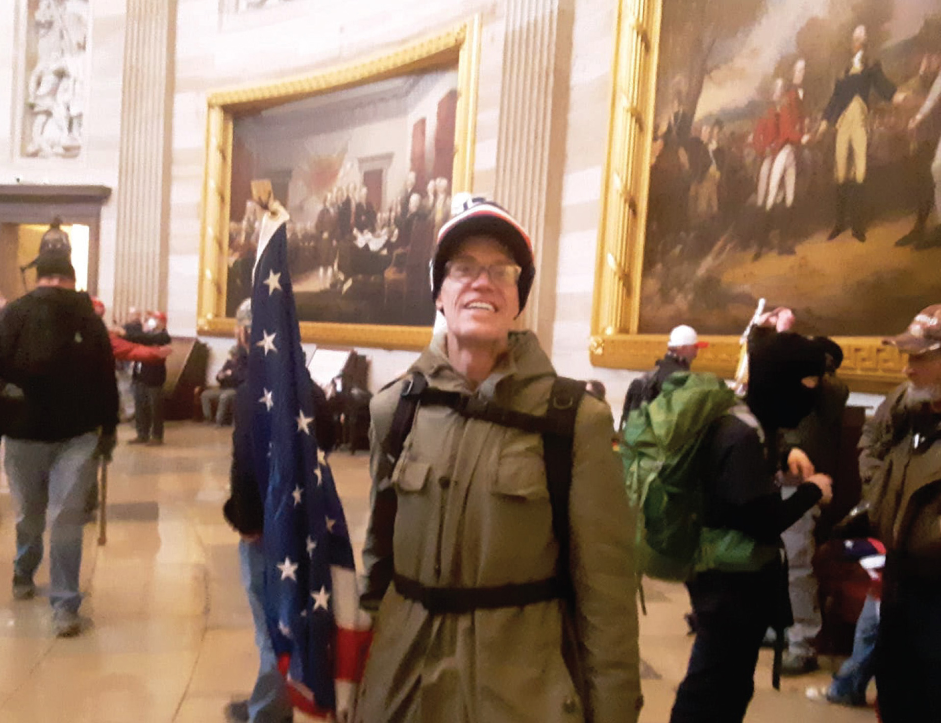 A person identified as Kevin Loftus poses for a photo while holding a U.S. flag while standing inside a room with curved marble masonry walls, a sculpture in a niche above a doorway and two visible paintings — "Declaration of Independence" by "Surrender of General Burgoyne" by John Trumbull — with other people standing and sitting in the background.