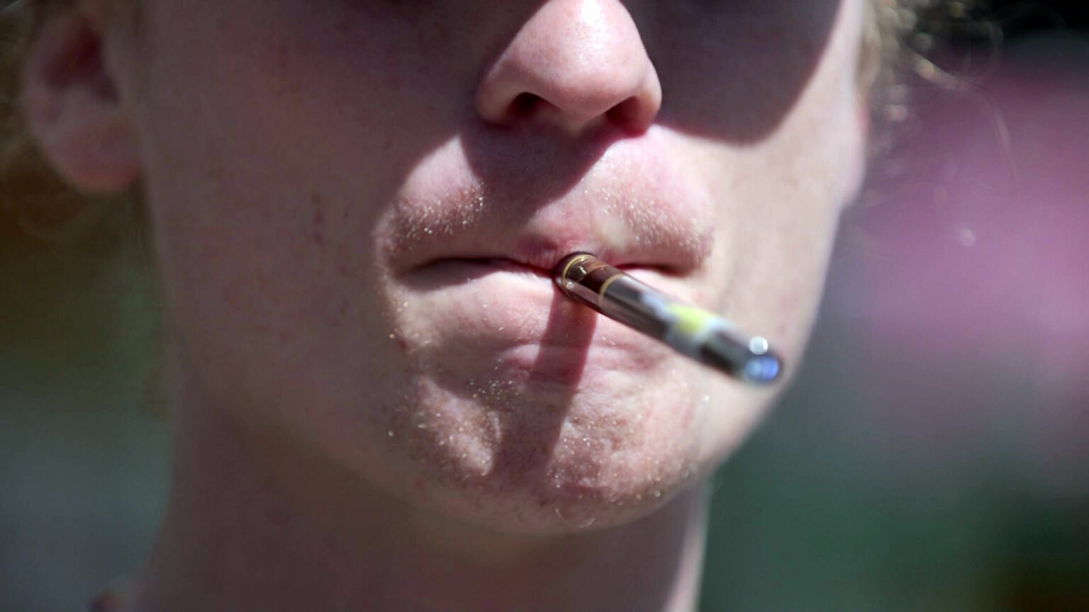 A closeup image shows the lower face of a man who is vaping on an e-cigarette.
