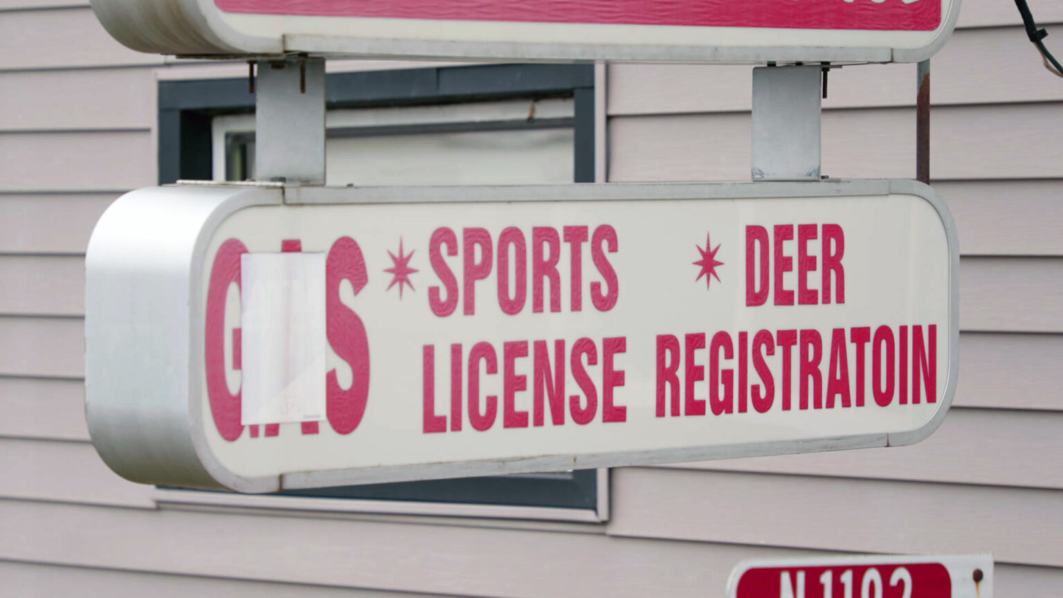 A sign with internal illumination that is not activated and the words Gas, which is covered in part by tape, as well as Sports License and Deer Registration, is mounted to the the exterior wall of a building with horizontal siding.