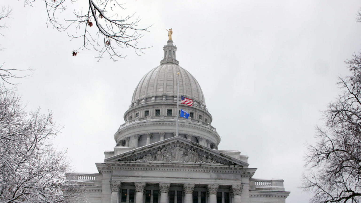 The U.S. and Wisconsin flags fly at half staff on a flagpole mounted to the top of a pediment on one wing of a marble masonry building with pillars, relief statuary and a dome topped by a statue, with snow-covered tree branches on either side and in the foreground, under an overcast sky.
