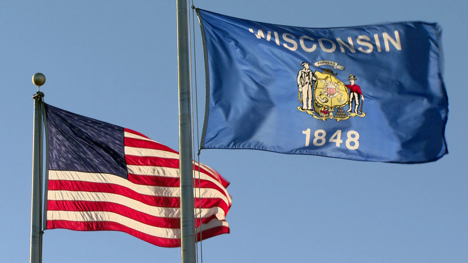 The U.S. and Wisconsin flags wave while attached to flagpoles, with a clear sky in the background.