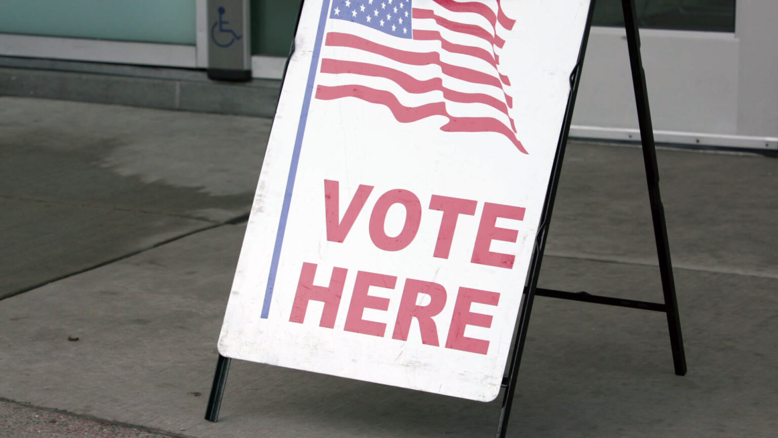 A sandwich board sign with a graphic of a U.S. flat and the words VOTE HERE stands on pavement in front of closed doors.