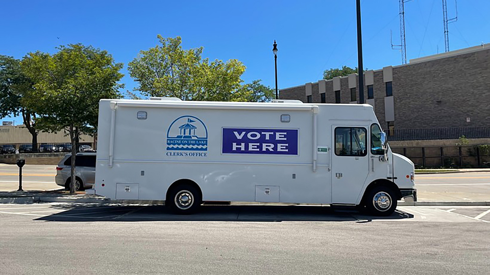 A step van with the logo of the City of Racine Clerk's office logo and the words VOTE HERE painted on its side is parked in a parking lot, with other vehicles, trees and a building in the background.
