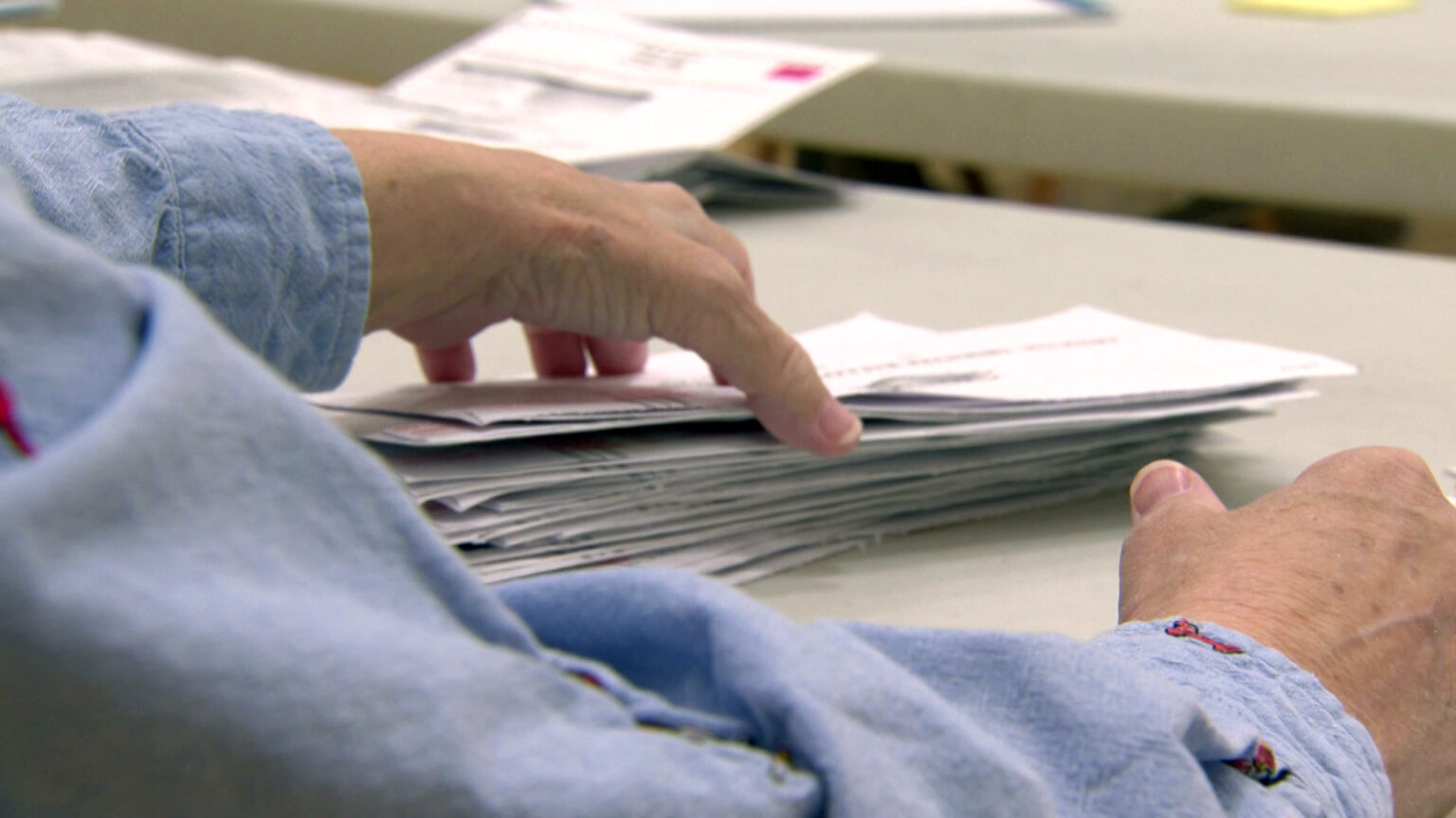 A poll worker's right hand sits on a the surface of a table while their left hand holds a stack of envelopes.