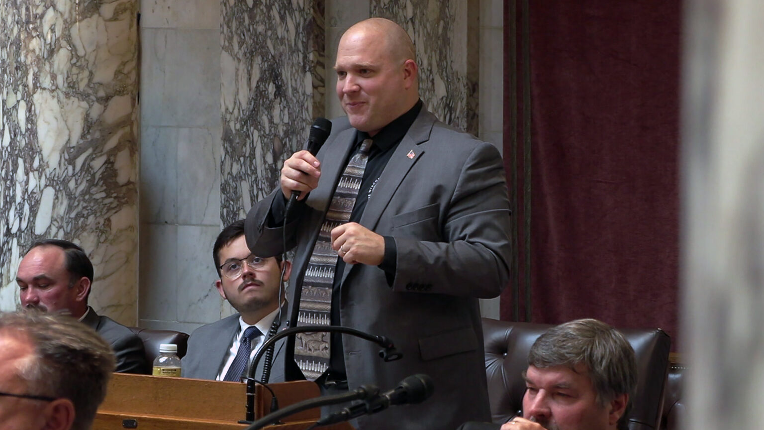 Shae Sortwell stands and speaks while holding a microphone in his right hand, with other people seated in front of and behind him, in a room with marble pillars and masonry.