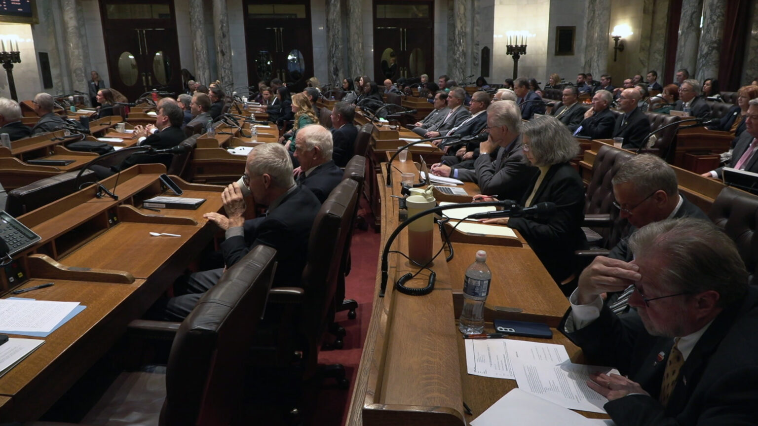 Wisconsin Assembly members sit in high-backed leather chairs on multiple rows of long wood desks arrayed in a semi circle, in a room with marble masonry and double doors with oval windows.